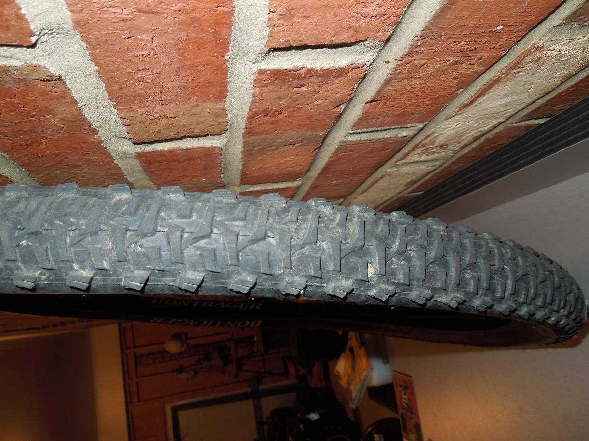 Bontrager 29-3: A close-up image of a bicycle tire mounted on a wall, showcasing its tread pattern. The background features a brick wall with a rough texture, and some tools and items are faintly visible in the lower part of the image.