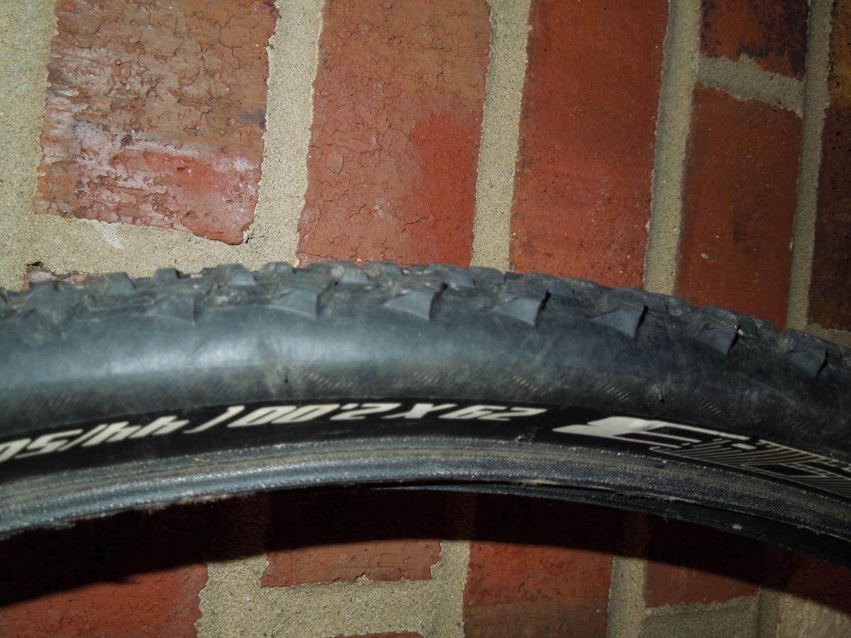 Bontrager 29-3: A close-up image of a bicycle tire resting against a textured brick wall. The tire features visible tread patterns and markings along the sidewall, with a focus on its detail. The brick wall, in various shades of red and brown, provides a contrasting background.