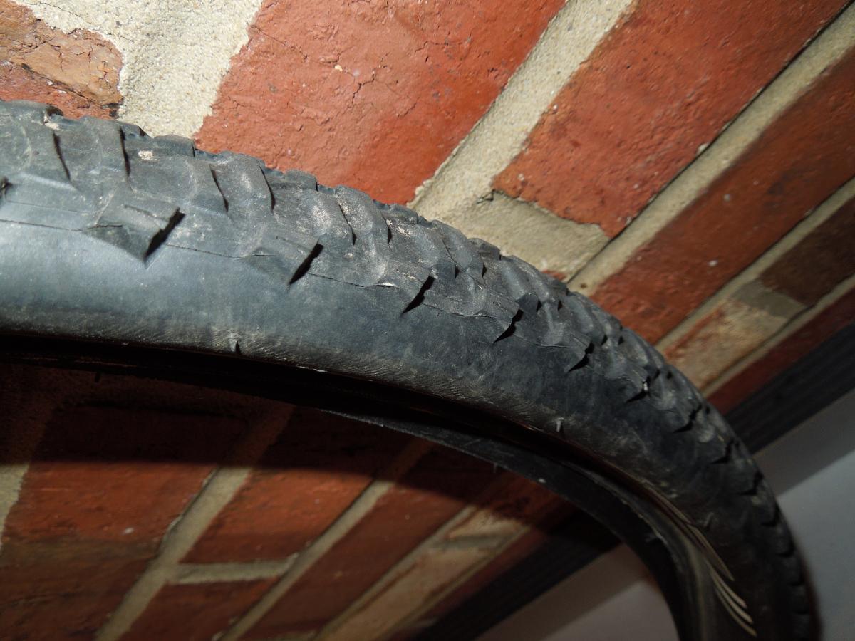 Bontrager 29-3: A close-up view of a bicycle tire showing its tread pattern, mounted against a brick wall. The tire appears used with visible wear, and the colors are predominantly black and gray, contrasting with the red and textured surface of the brick behind it.