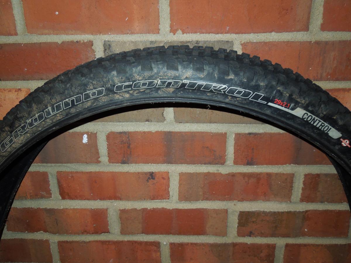 Specialized Ground Control: A close-up image of a bicycle tire mounted on a wall made of red bricks. The tire features the text "Ground Control" prominently displayed on its sidewall, along with additional specifications indicating it is a 29x2.1 model. The tread pattern of the tire is visible, showing signs of use and dirt.