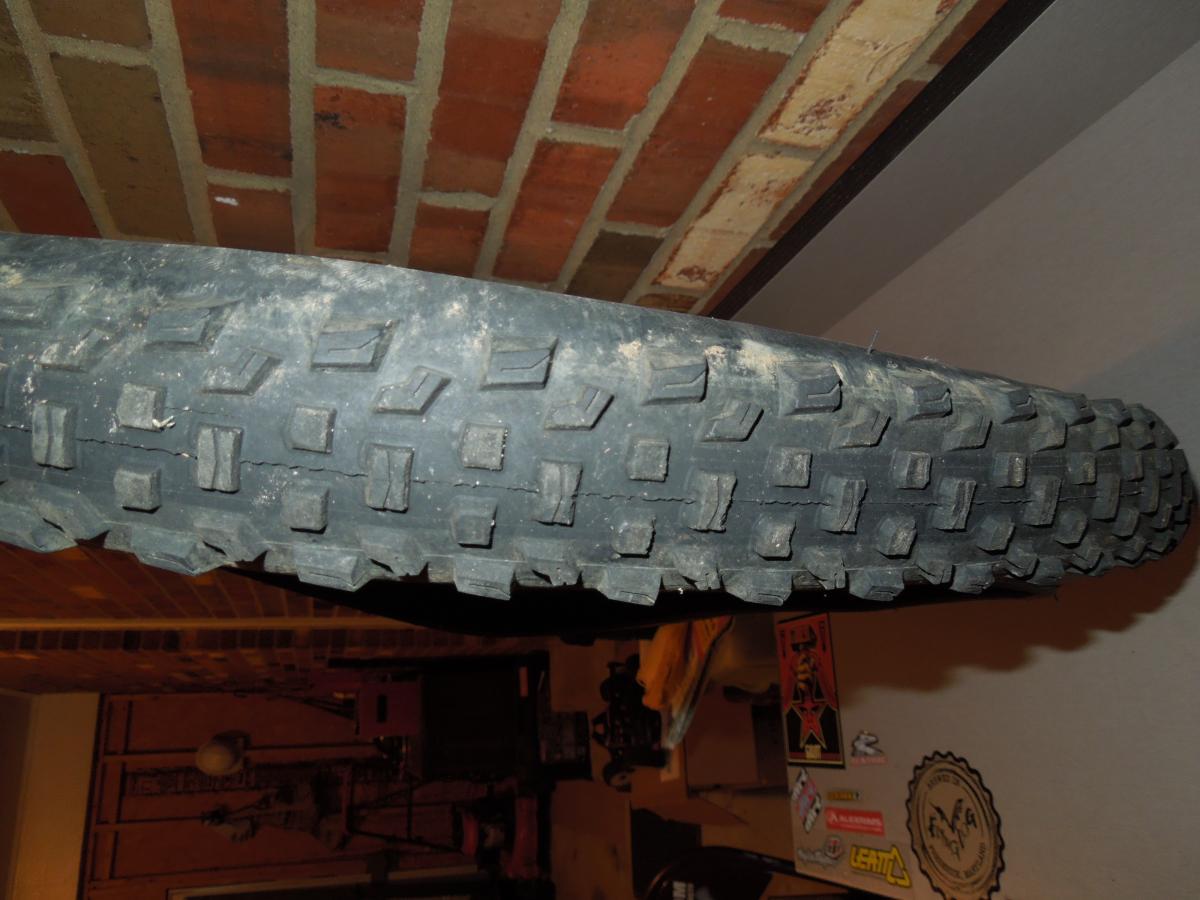 Specialized Ground Control: Close-up image of a dirt bike tire with a rugged tread pattern, resting against a background of brick wall and various stickers and objects on a surface below.