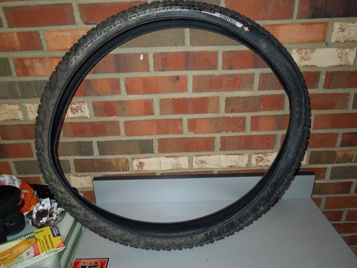 Specialized Ground Control: A close-up image of a mountain bike tire mounted on a table, showcasing the tire's tread pattern and sidewall. The background features a brick wall and various items on a countertop, including crumpled papers and a black container. The tire appears slightly dirty, indicating usage.