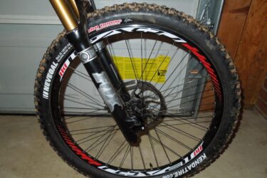 Kenda Nevegal John Tomac Signature Series: Close-up view of a mountain bike front wheel and fork, showcasing a muddy tire with the "Kenda" branding, a black and silver FOX suspension fork, and a visible disc brake. The background features a garage setting with a partially open door and a warning label on the wall.