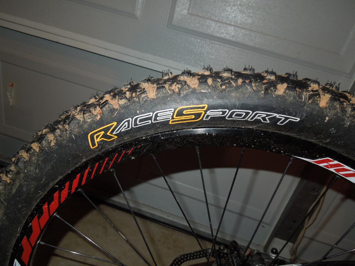 Continental Mountain King Race Sport: Close-up image of a mountain bike tire with a tan and black tread pattern. The tire has the brand name "RaceSport" prominently displayed in white and orange letters. The background features a gray garage door and part of the bike frame. The tire shows signs of use with some dirt and debris on the tread.