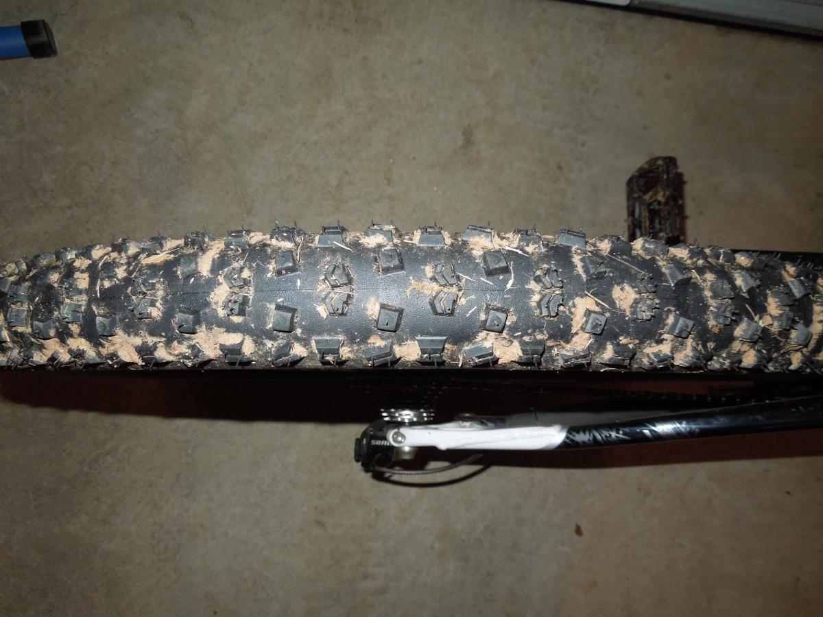 Continental Mountain King Race Sport: Close-up of a muddy mountain bike tire, showing a detailed tread pattern. The tire is partially visible, resting on a concrete surface with dirt and mud caked on it. The bike frame and components are also faintly visible in the background.