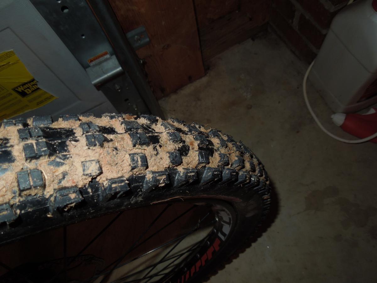 Kenda Nevegal John Tomac Signature Series: Close-up view of a muddy bike tire with a knobby tread pattern, set against a concrete floor background. The tire shows signs of recent off-road use, with dirt and debris clinging to the surface. In the background, a partially visible wall and a red container are present.