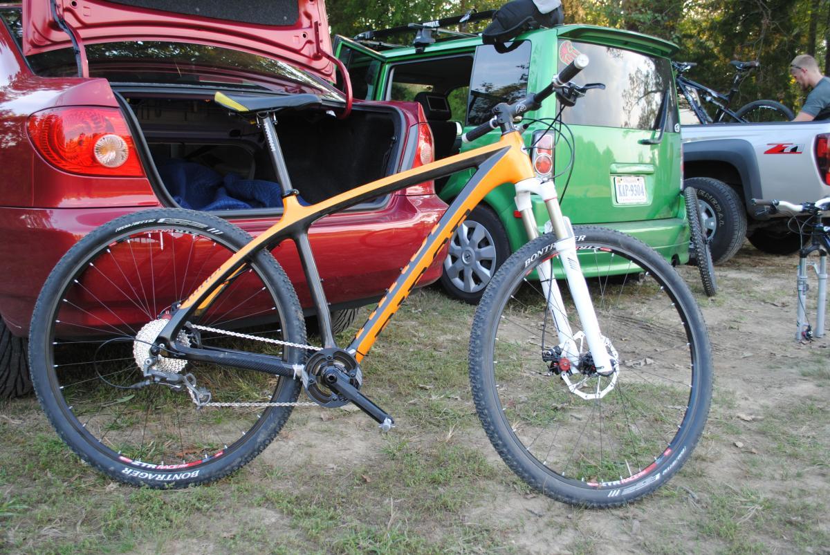 Niner Air 9 Carbon: An orange mountain bike stands upright on grass, partially in view of a maroon car's open trunk, which reveals a blanket inside. A green vehicle is parked nearby, and there are additional bikes and people in the background, suggesting a recreational outdoor setting.