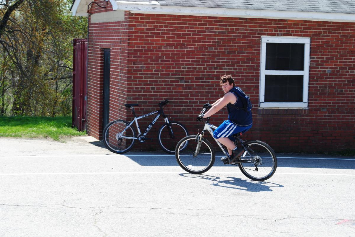 Trek 4300: A young person wearing a sleeveless blue shirt and blue athletic shorts rides a bicycle on a paved surface near a red brick building. A second bike is parked against the building in the background. The scene is outdoors on a sunny day.