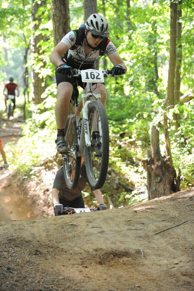 On-One Scandal 29er Vertical Dropouts: A mountain biker, wearing a helmet and race number 162, performs a jump over a dirt ramp on a wooded trail. In the background, another cyclist can be seen riding along the path. Sunlight filters through the trees, creating a vibrant green backdrop.
