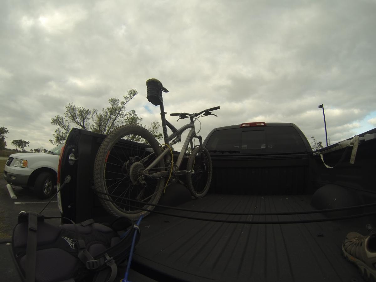 Specialized Camber: A mountain bike secured in the bed of a pickup truck, with a cloudy sky in the background. A harness and a pair of shoes are visible on the truck's floor. Nearby, another vehicle can be seen in the parking lot.