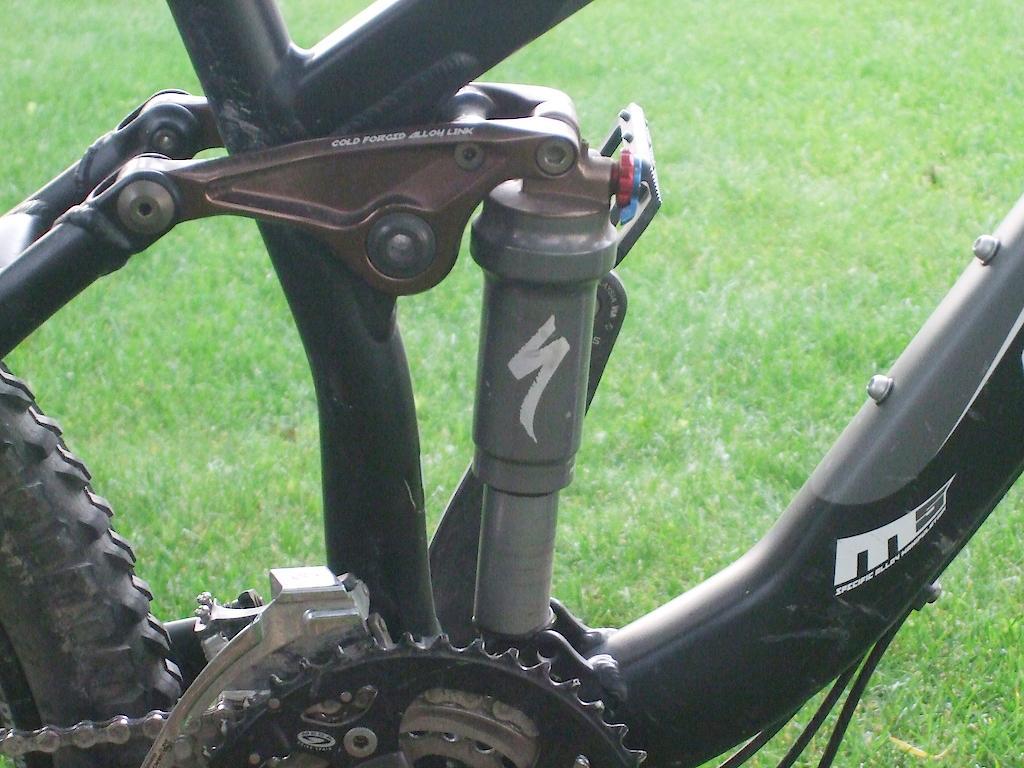 Specialized Enduro SL Comp FSR: Detailed close-up of a mountain bike's rear suspension system, highlighting the shock absorber and linkage components. The image shows a black bike frame with technical features, set against a green grass background.