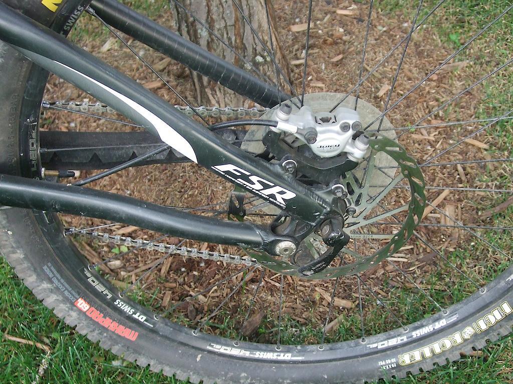 Specialized Enduro SL Comp FSR: A close-up view of a bicycle's rear wheel and drivetrain, showcasing the disc brake, gear mechanism, and portion of the tire on grass and mulch background.