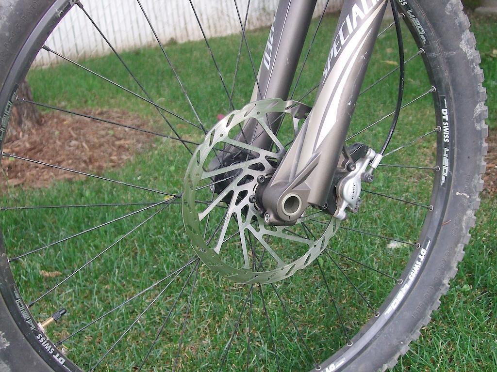 Specialized Enduro SL Comp FSR: Close-up view of a mountain bike's front wheel, showcasing a metal disc brake attached to a fork, with green grass in the background. The wheel features a rugged tire suitable for off-road biking.