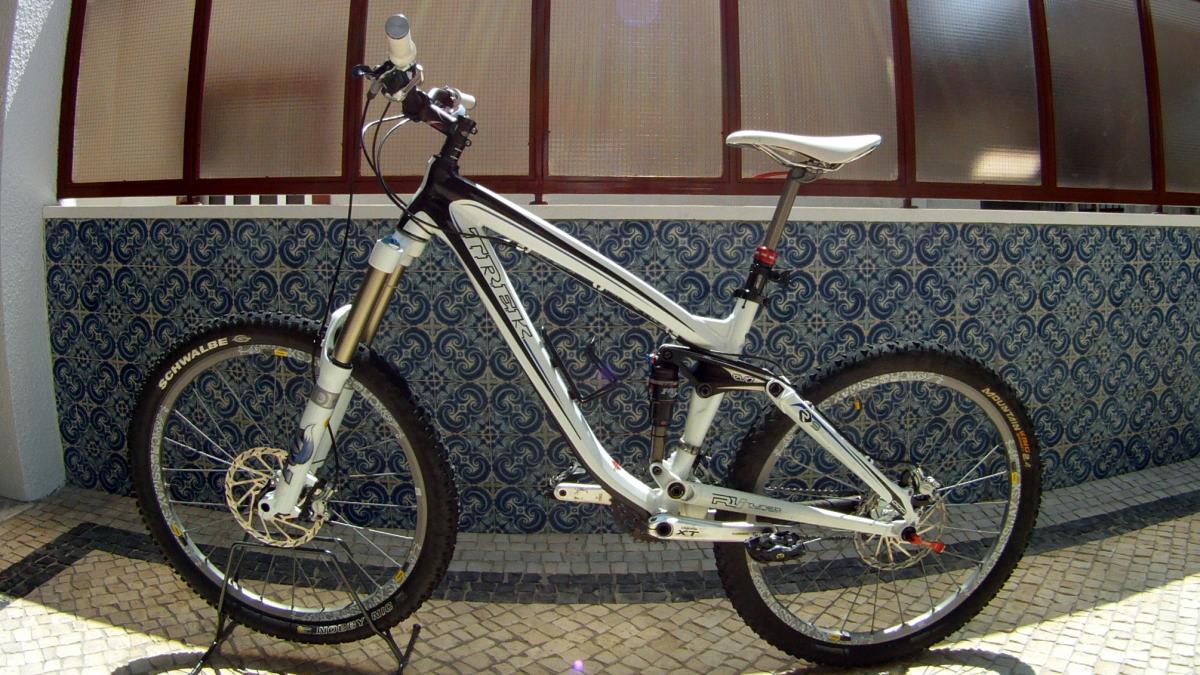 Trek Remedy 9: A white and black mountain bike is displayed upright on a stand. It features a modern design with front suspension and thick, rugged tires, suitable for off-road terrain. The backdrop includes a patterned blue and white wall, with light streaming in from a window, highlighting the bike's details.
