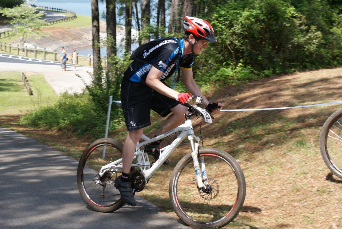 Airborne Zeppelin Elite: A person riding a mountain bike on a dirt path surrounded by trees, wearing a black and blue cycling jersey and red gloves. The background features a scenic view of a water body and a winding road.