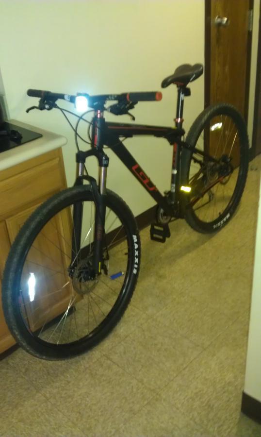GT Karakoram: A black mountain bike with red accents is parked inside a room, next to a wooden kitchen cabinet. The bike features wide tires marked with "Maxxis" and has a headlight attached to the handlebars. The room has a beige floor and a wooden door in the background.