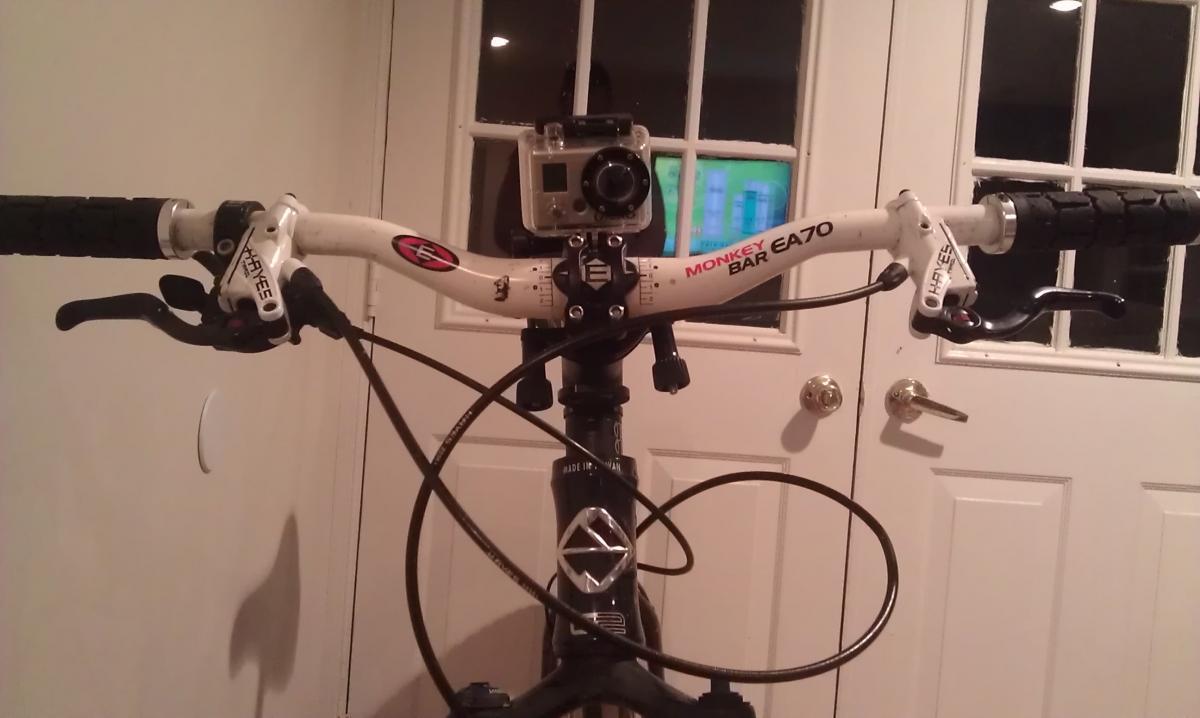 GoPro Roll Bar Mount: Close-up view of a bicycle handlebar featuring a GoPro camera mounted on top. The handlebar has black grips and two brake levers, with cables visible. In the background, there is a glimpse of a door and a window. The image is set indoors with soft lighting.