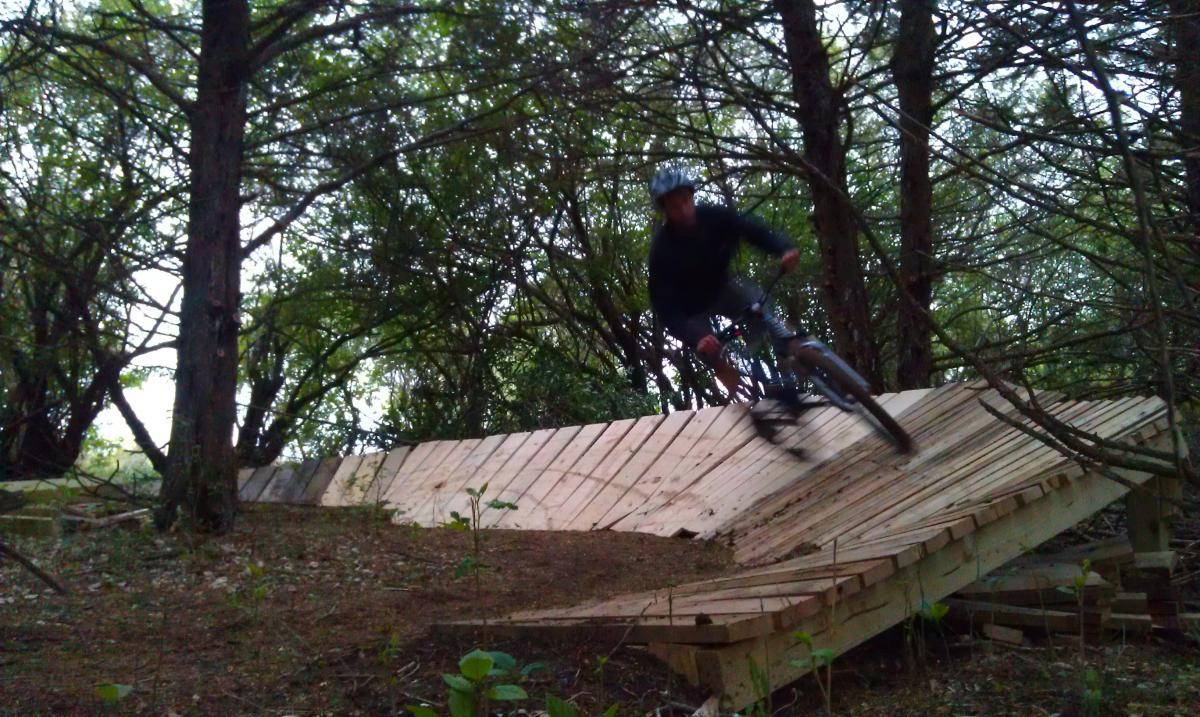 A mountain biker rides on an incline made of wooden planks, surrounded by trees in a wooded area. The biker is in motion, captured in a dynamic pose, emphasizing speed and action. Maskenthine Lake mountain bike trail.