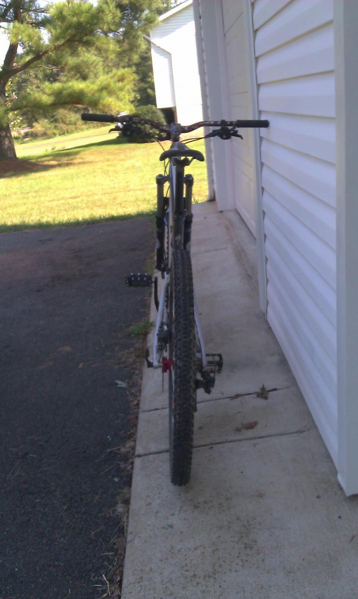 Specialized Stumpjumper FSR Comp 29: Mountain bike leaning against a white wall, with the handlebars and rear wheel visible. The background features a grassy area and some trees.