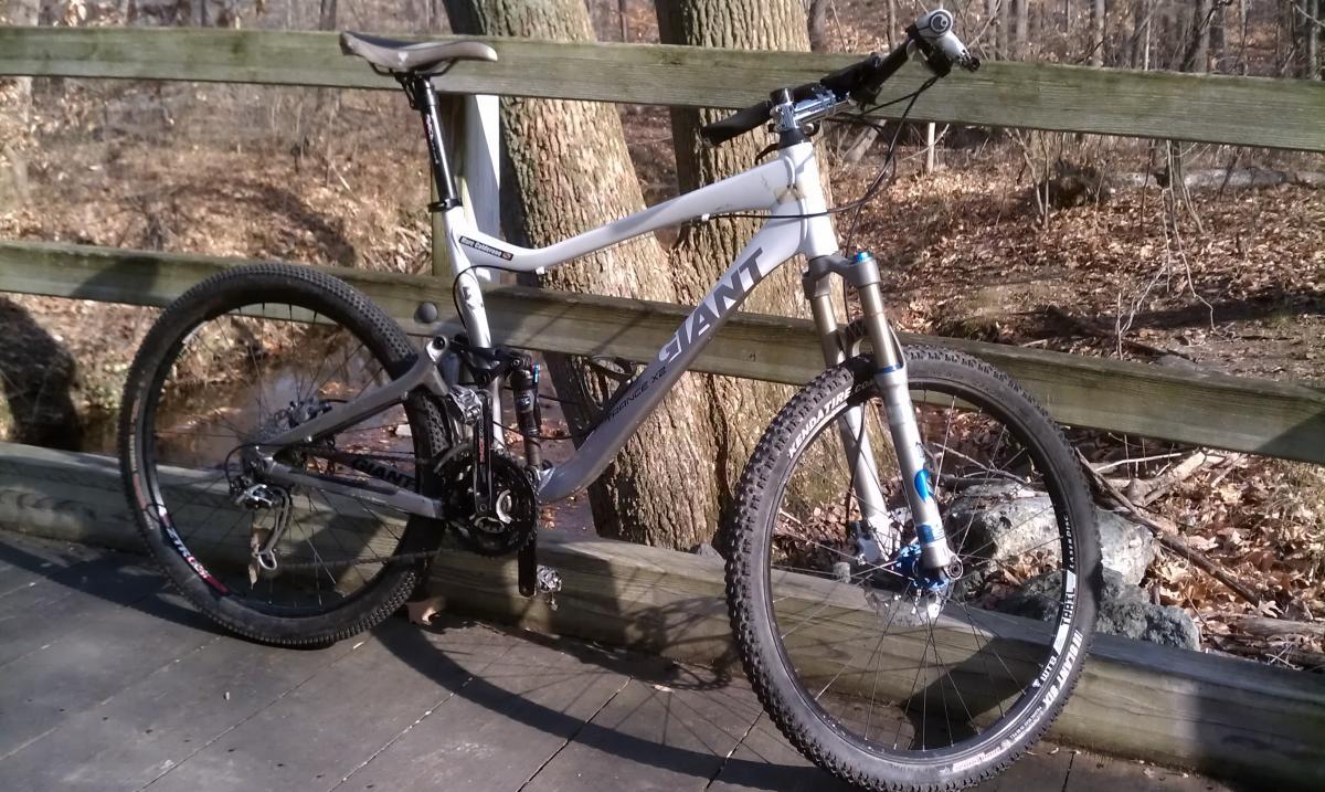 Giant Trance X2: A gray mountain bike leaning against a wooden railing in a wooded area, with trees in the background and a hint of a stream visible. The bike features thick tires and a sturdy frame, ideal for off-road trails.