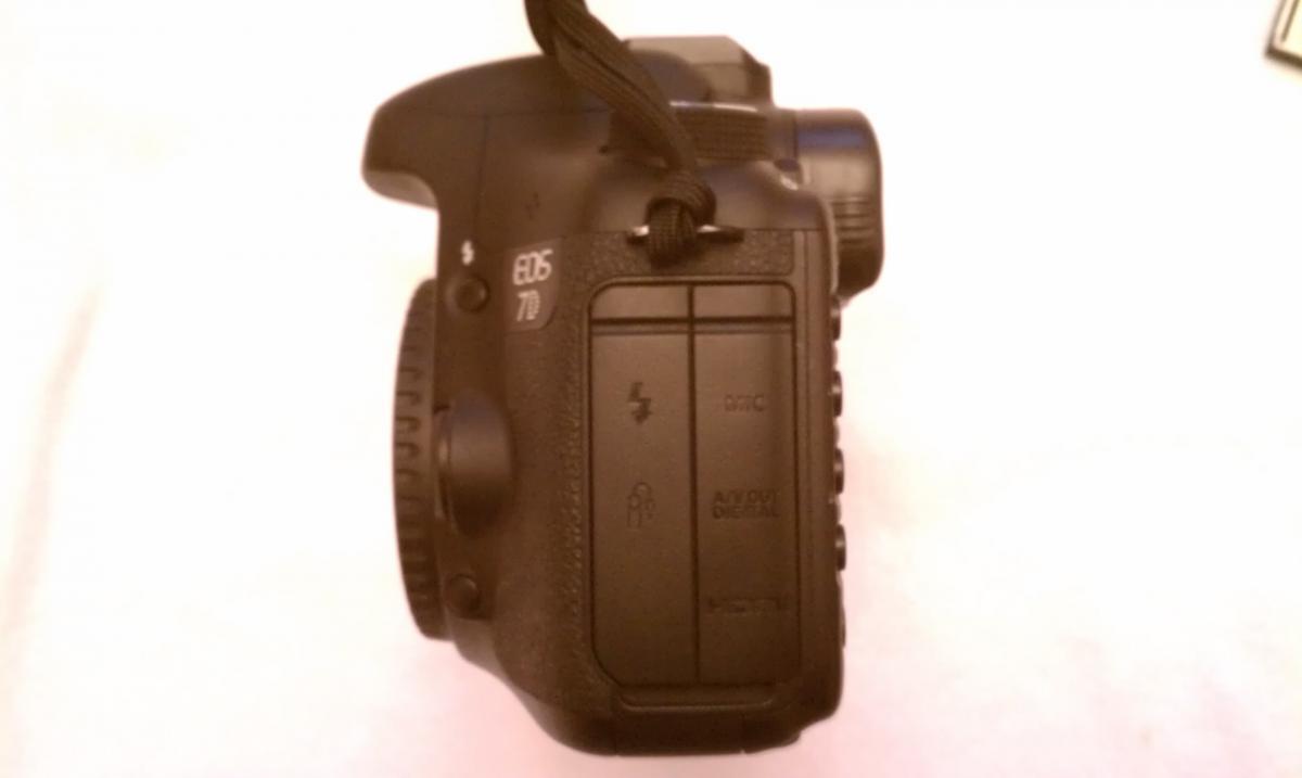Canon 7D: A close-up view of the side of a DSLR camera, showing various buttons and ports. The camera is predominantly black with a textured grip. There are labels indicating settings and connections, including a flash symbol and HDMI port. A strap is attached for easy handling. The background is a plain white surface.