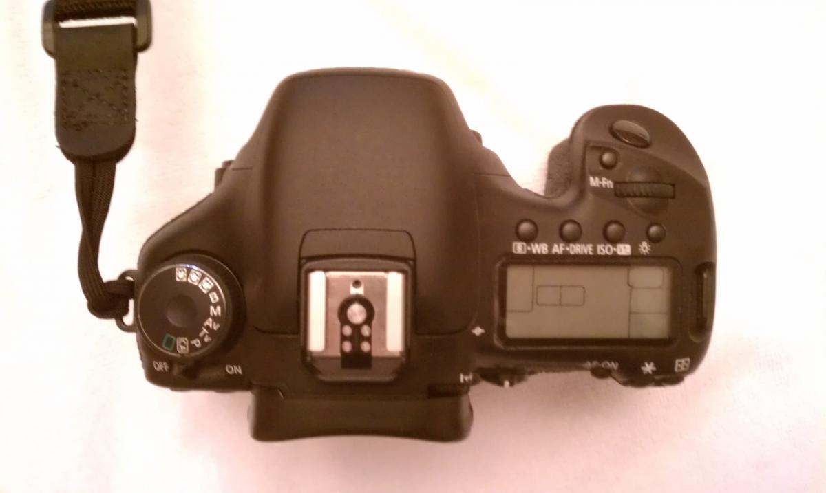 Canon 7D: A top view of a digital camera featuring various dials, buttons, and an LCD display, with a strap attached on the left side. The camera is positioned on a light-colored background.