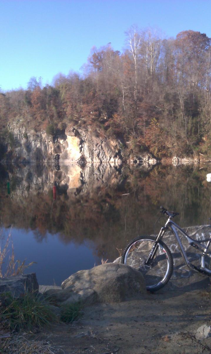 Specialized Stumpjumper FSR Comp 29: A mountain bike rests on a rocky shore beside a calm lake, surrounded by trees displaying autumn colors. The reflection of a rocky cliff and colorful markers can be seen in the water, under a clear blue sky.