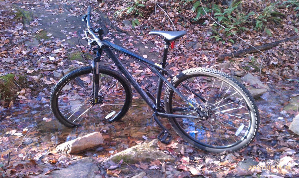 GT Karakoram: A black mountain bike stands in a shallow stream surrounded by fallen leaves and rocky terrain, with greenery in the background.