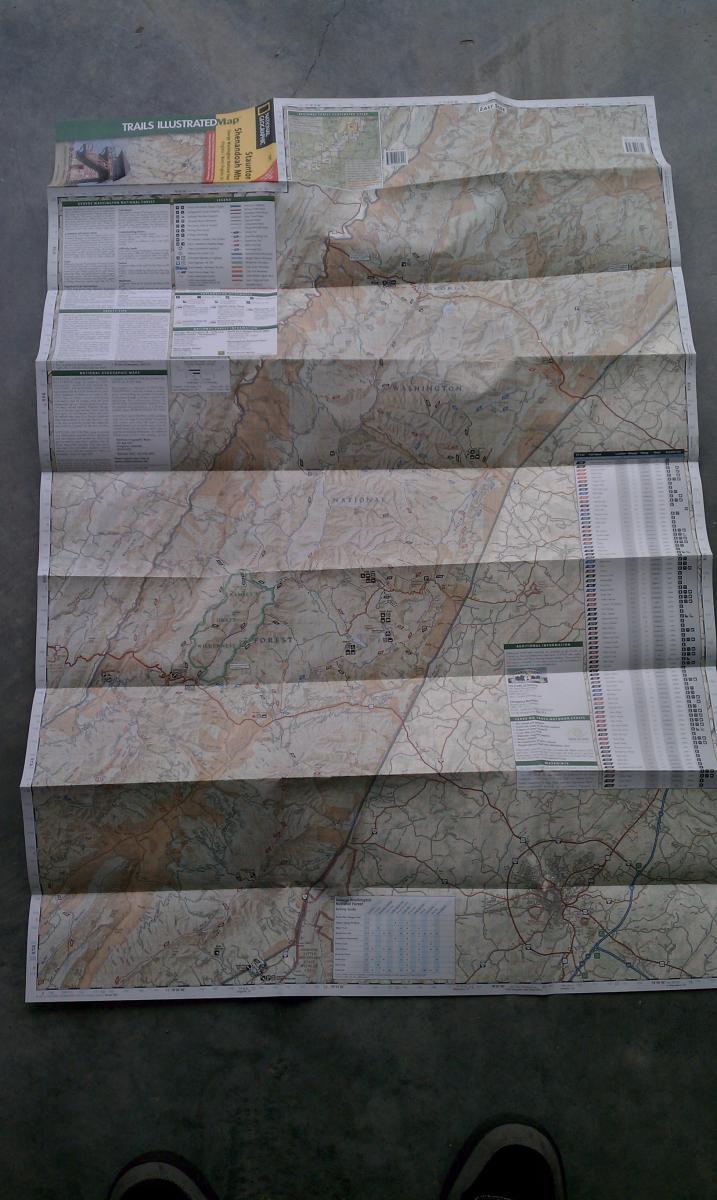 National Geographic Trails Illustrated #791 Staunton Shenandoah Mountain: A folded Trails Illustrated map spread out on a flat surface, showing topographical features, trails, and roads in a natural area. The map includes informational sections, icons indicating points of interest, and a legend. The bottom of the image shows a pair of shoes.