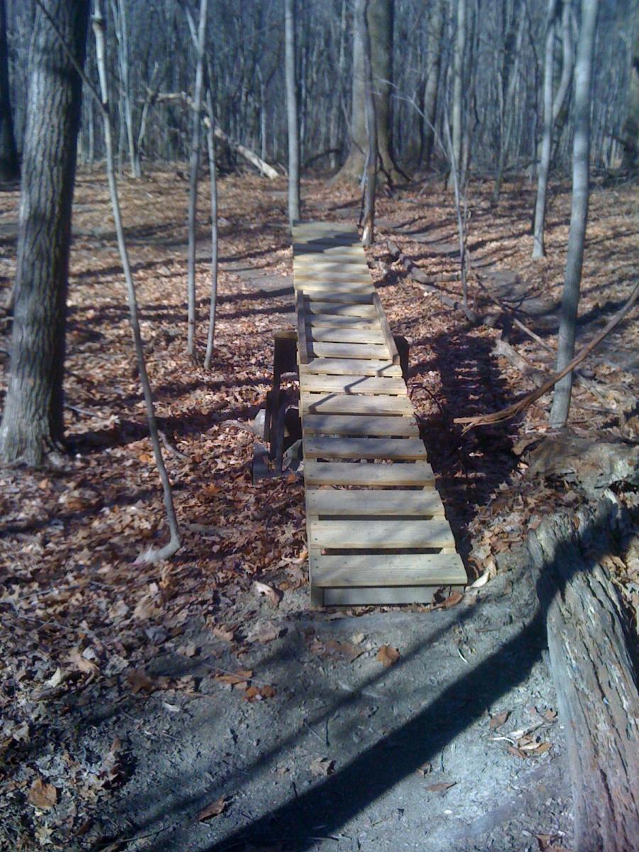 A wooden bridge spanning a small gap in a forested area, surrounded by bare trees and scattered brown leaves on the ground. The bridge is made of planks and sits on supportive structures, leading into the trees on the other side. Bakers Ridge mountain bike trail.