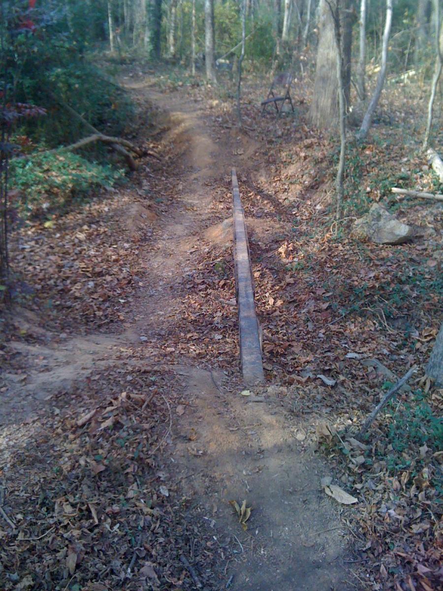 A narrow dirt path in a wooded area, with a wooden plank serving as a bridge over a small dip in the trail. The ground is covered with fallen leaves, and a weathered chair can be seen to the side among the trees. Back Yard Trails mountain bike trail.
