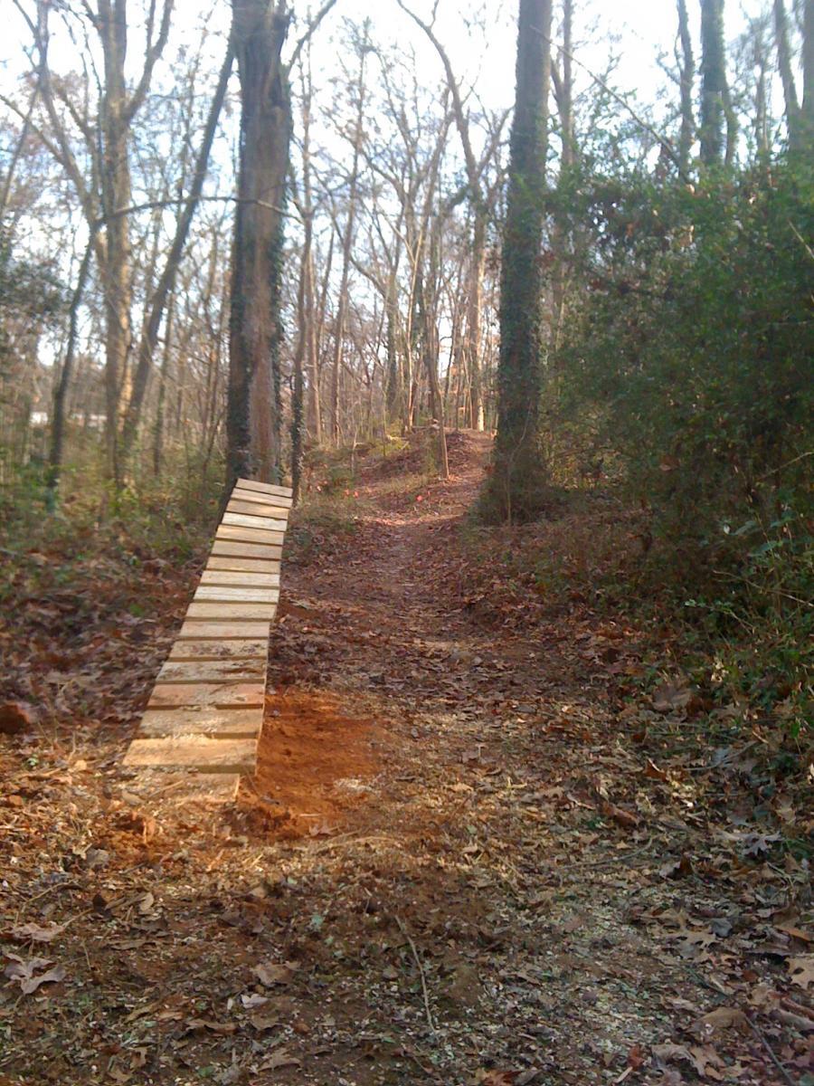 A wooden ramp positioned on a dirt trail surrounded by trees in a forest setting, with fallen leaves and patches of bare ground visible. The ramp leads up an incline in the path, suggesting a mountain biking or hiking route through the natural landscape. Back Yard Trails mountain bike trail.