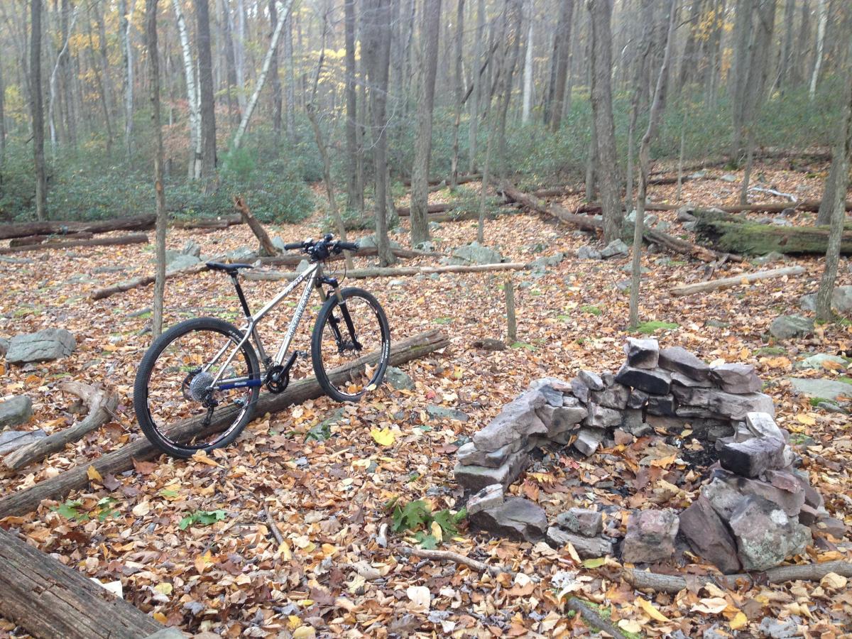 Motobecane Fly Team 29er Ti X0 Edition: A mountain bike parked on a trail surrounded by autumn foliage in a forest. In the foreground, a circular stone fire pit is visible, with fallen leaves covering the ground and trees in the background.