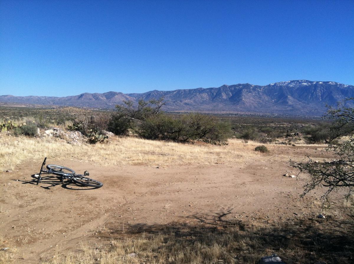 A mountain bike lying on its side in a dry, open landscape with sparse vegetation and rocky soil. In the background, there are rolling hills and mountains under a clear blue sky. The scene captures a sense of tranquility and rugged natural beauty. Honeybee Canyon / Rail X Ranch mountain bike trail.