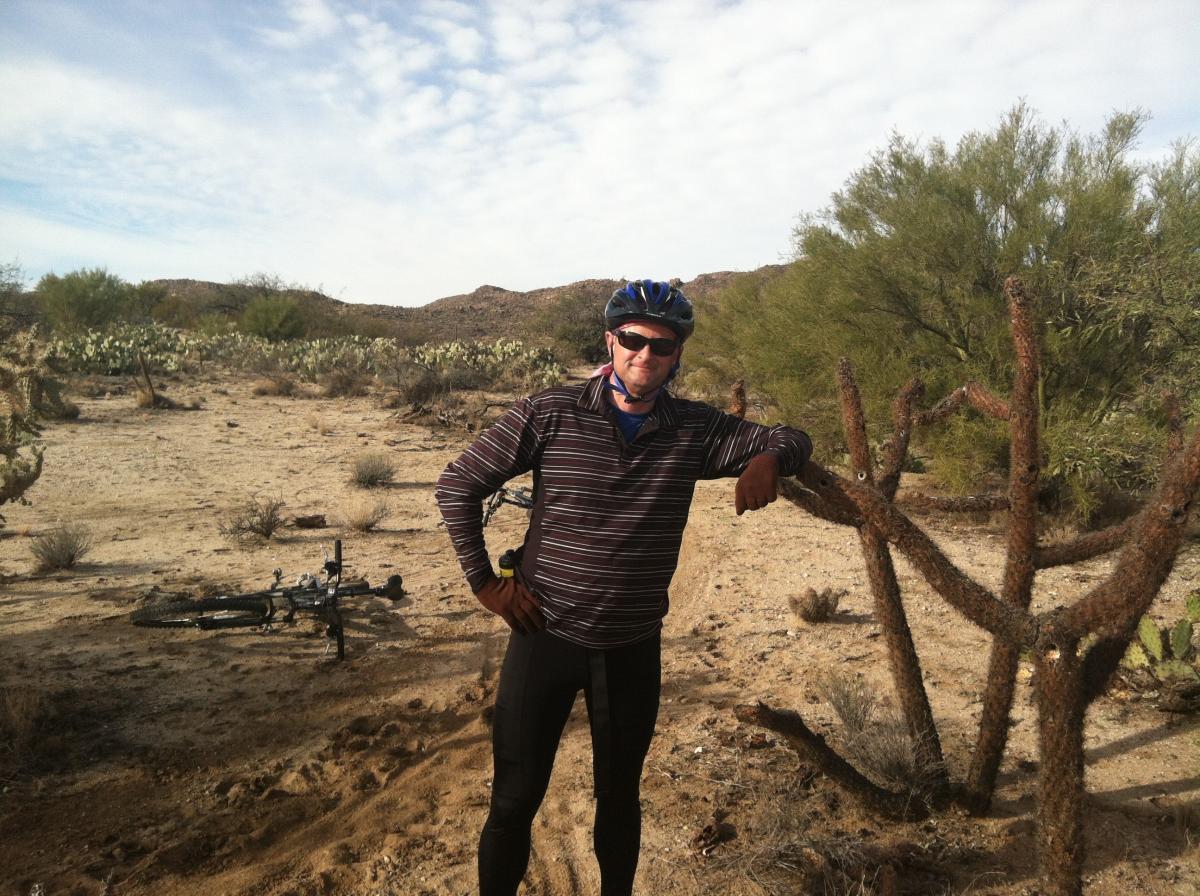 A man wearing a helmet and sunglasses stands next to a cactus in a desert landscape, with a mountain range in the background. A mountain bike is leaning on the ground nearby. The sky is partly cloudy, and the surroundings feature sparse vegetation typical of a desert environment. Honeybee Canyon / Rail X Ranch mountain bike trail.