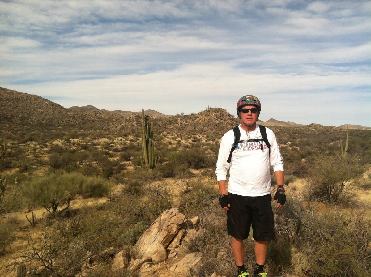 A person wearing sunglasses, a helmet, and a long-sleeve shirt stands in a desert landscape. The background features mountains and various desert vegetation, including cacti and bushes, under a partly cloudy sky. Honeybee Canyon / Rail X Ranch mountain bike trail.