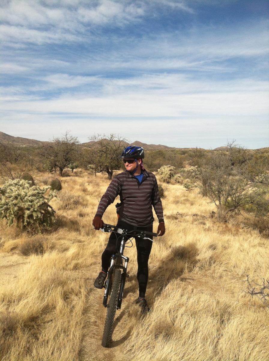 A person in athletic attire smiles while standing next to a mountain bike on a dirt trail surrounded by dry grass and sparse vegetation. The landscape features low bushes and distant hills under a partly cloudy sky. Honeybee Canyon / Rail X Ranch mountain bike trail.