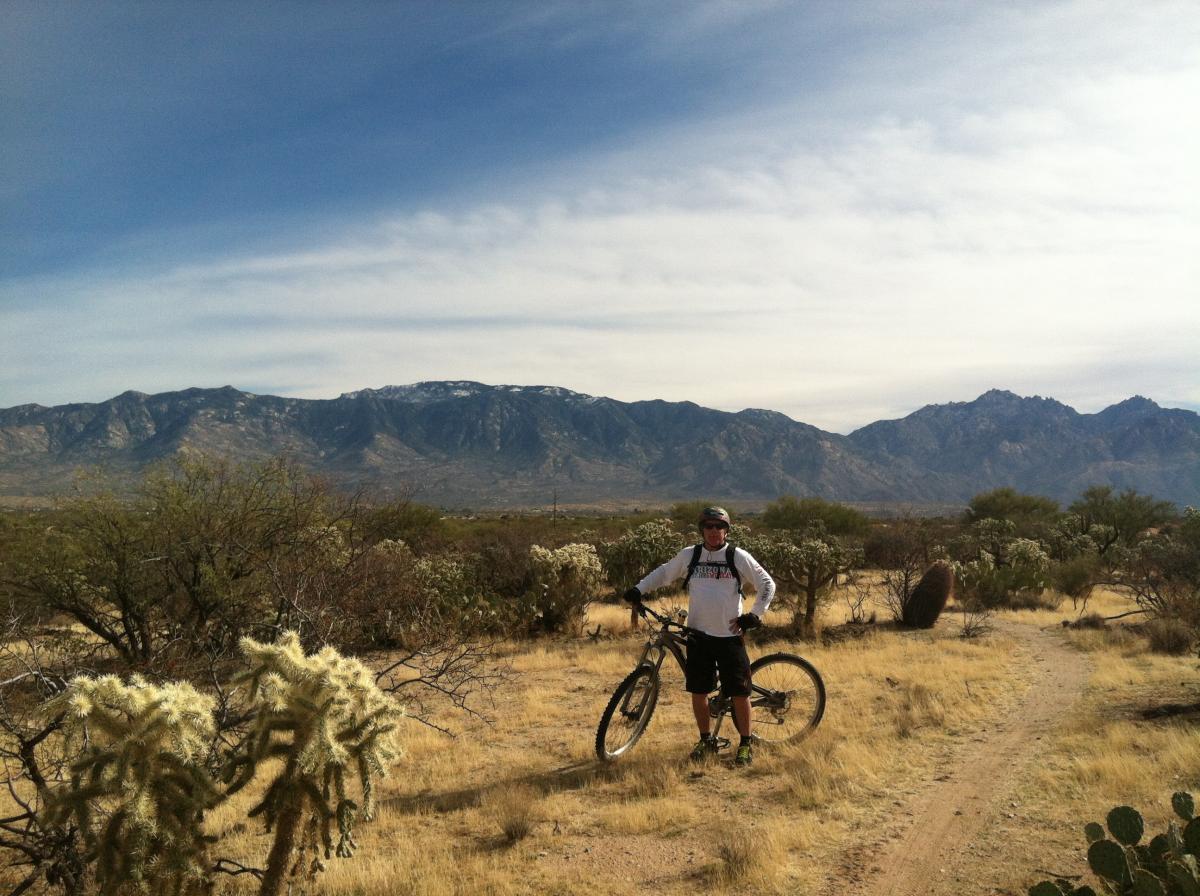 A mountain biker standing next to a bicycle on a dirt trail, surrounded by desert vegetation and cacti, with a mountainous landscape in the background under a clear sky. Honeybee Canyon / Rail X Ranch mountain bike trail.