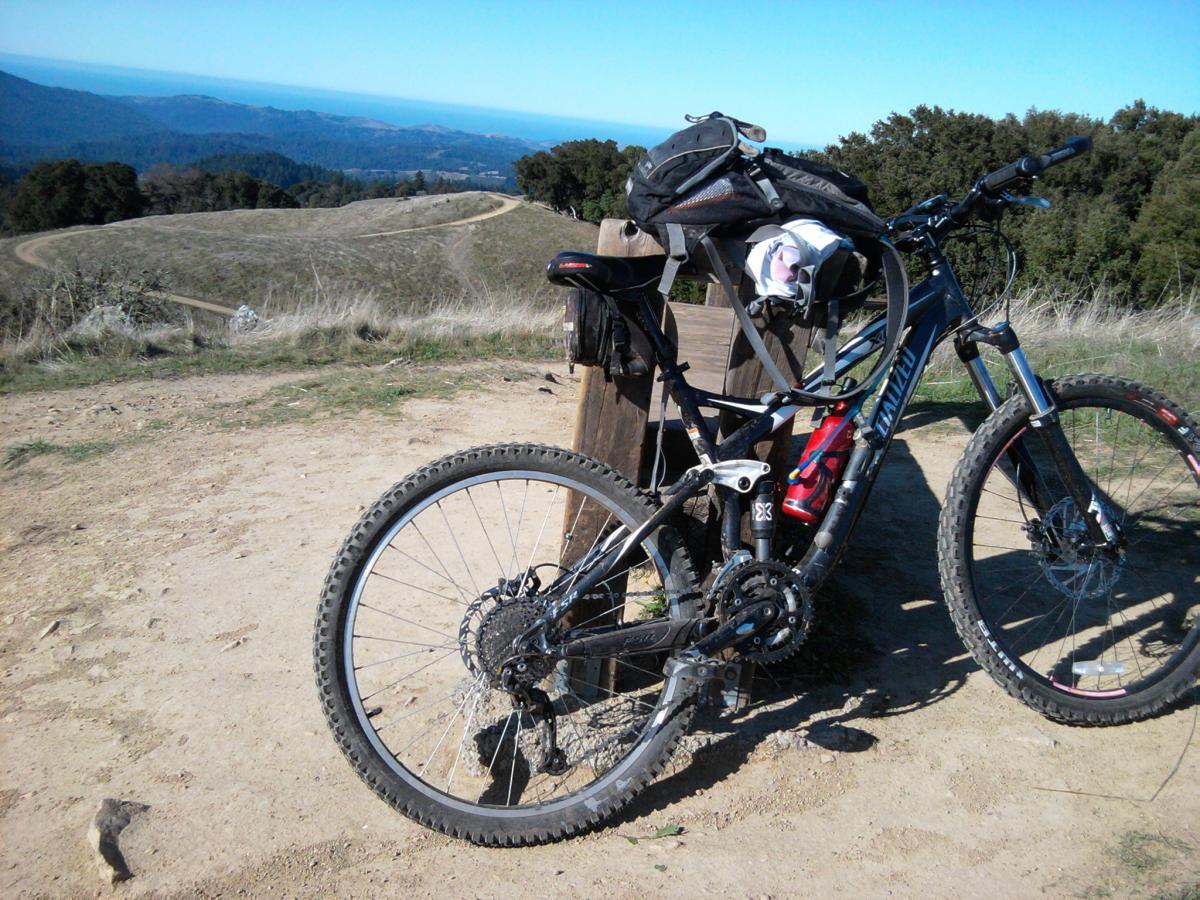 Specialized FSR XC Comp: A mountain bike parked beside a wooden bench on a dirt path, with a scenic view of rolling hills and the ocean in the background. The bike is equipped with a backpack and a water bottle, set against a clear blue sky.