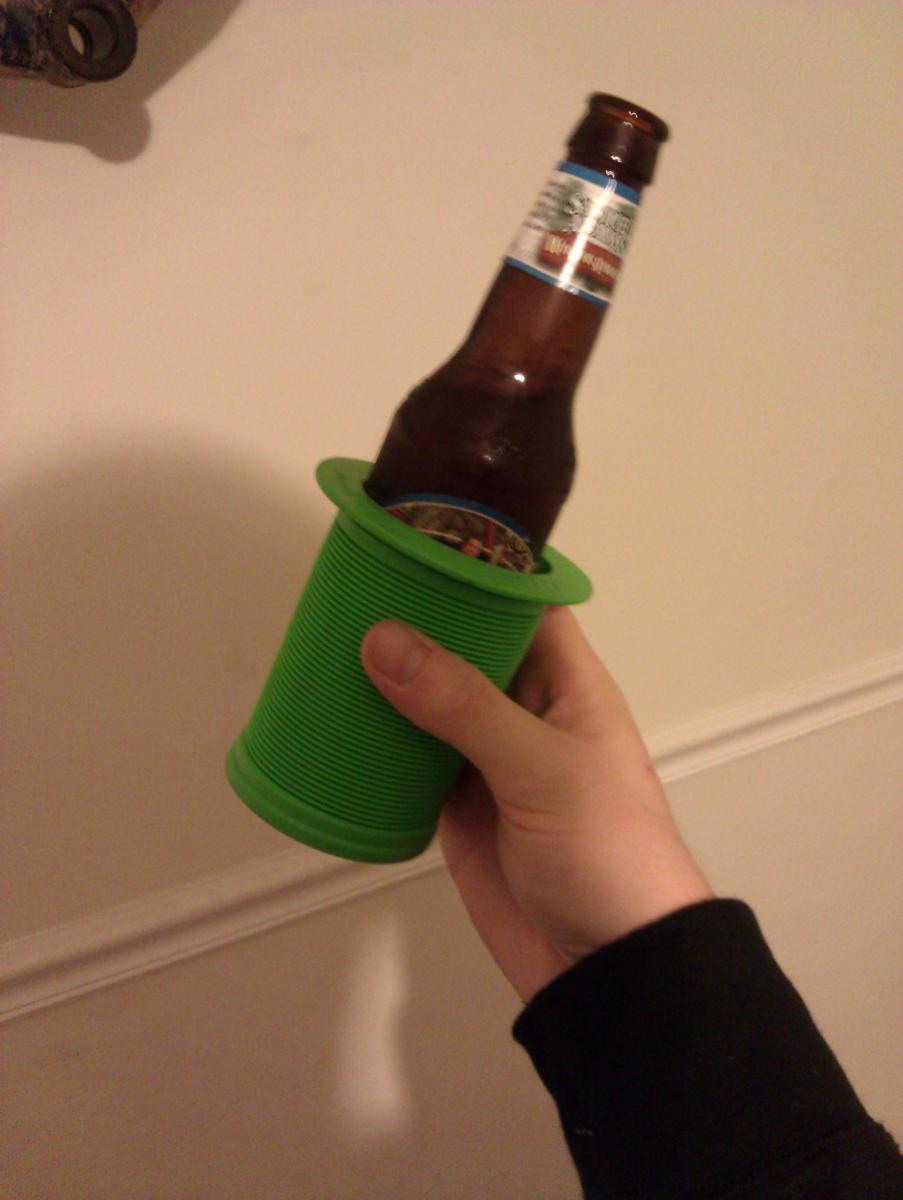 ODI ODI Koozie: A hand holding a green, ribbed bottle holder with a brown beer bottle inside, set against a plain wall.