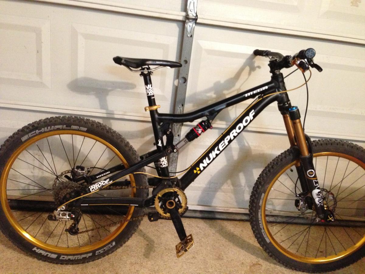 Nukeproof Mega: A black and gold mountain bike is leaning against a garage wall. The bike features wide tires with knobby tread, disc brakes, and a prominent "Nukeproof" logo on the frame. The handlebars are equipped with grips and a gear shifter. The background shows the garage door partially closed.