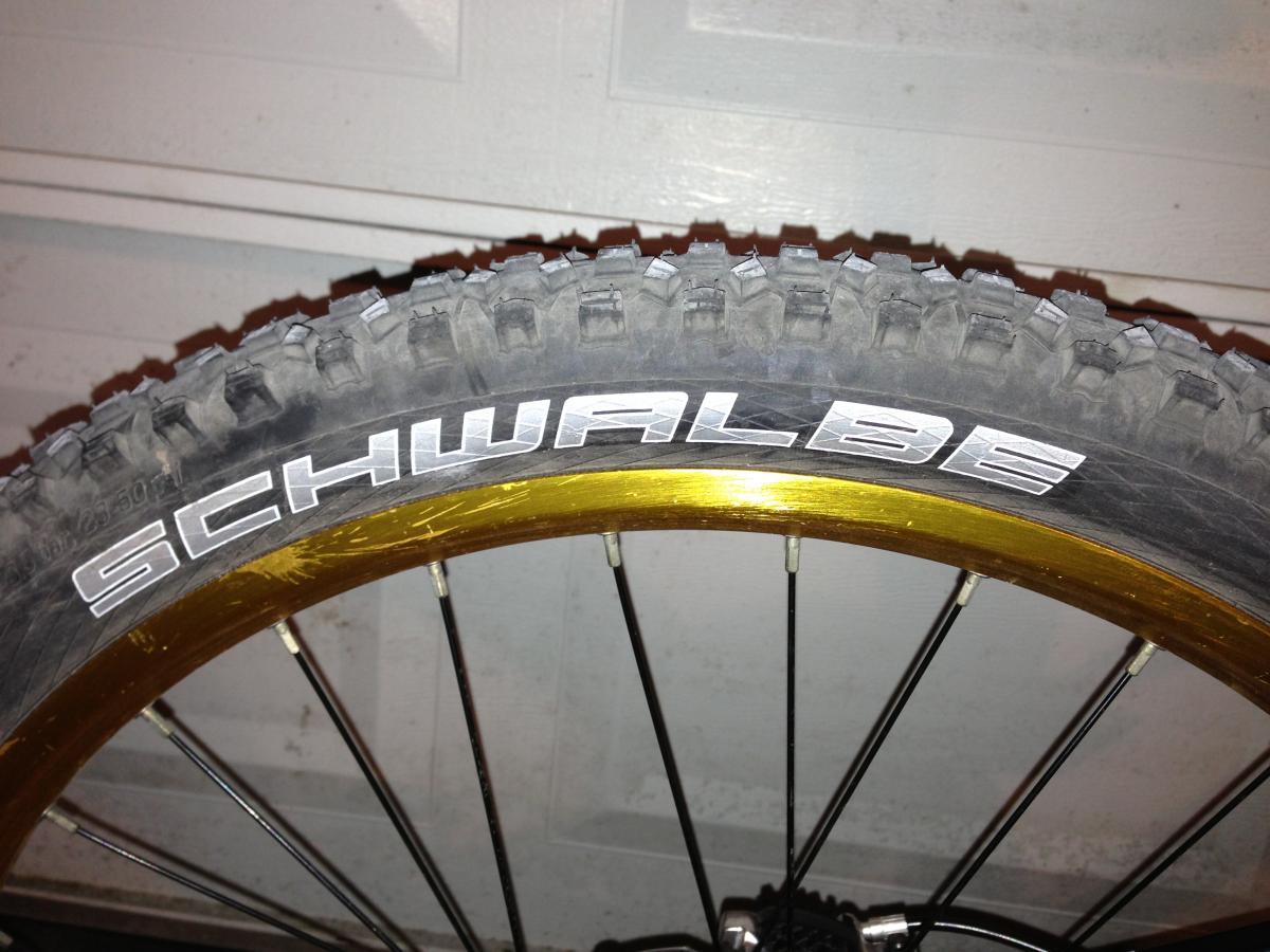 Schwalbe Hans Dampf: Close-up of a bicycle tire with the brand name "Schwalbe" displayed prominently in white letters on the sidewall. The tire features a rugged tread pattern designed for off-road use, and it is mounted on a gold-colored rim. The background is a plain garage wall.