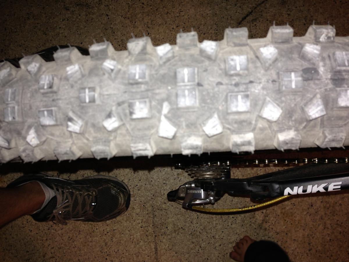 Schwalbe Hans Dampf: Close-up view of a mountain bike tire tread, showing clear patterns designed for traction, alongside a person's foot wearing a sneaker. The image also features part of the bike's chain and pedal. The setting appears to be indoors on a surface that resembles concrete.