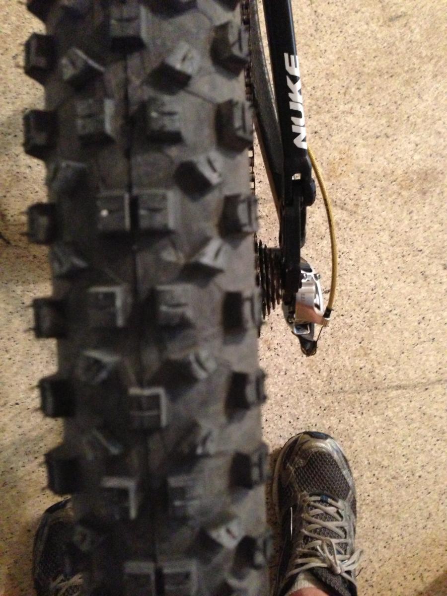 Schwalbe Hans Dampf: A close-up view of a mountain bike tire with deep, knobby tread, resting on a flat surface. In the foreground, a person’s foot in a gray athletic shoe is partially visible. The bike's frame and gearing components can be seen in the background.