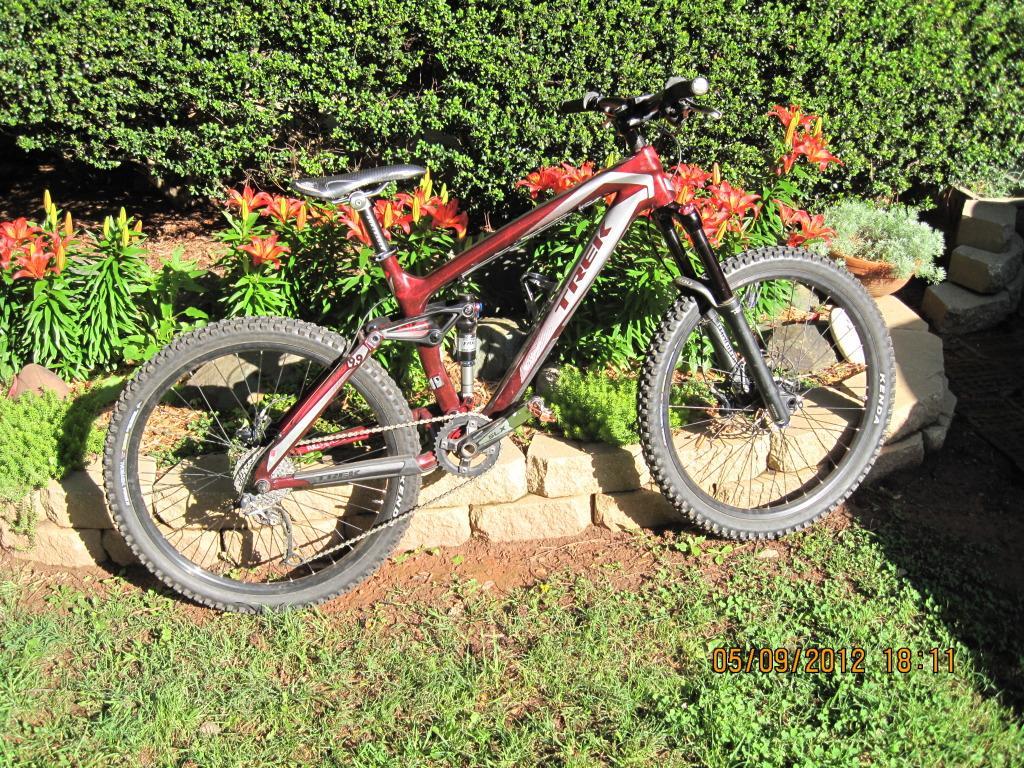 Trek Remedy 9.9: A red Trek mountain bike positioned on a grassy area, with a backdrop of colorful flowers and lush greenery. The bike features thick tires and a sturdy frame, suitable for off-road biking. Sunlight illuminates the scene, highlighting the bike's details.
