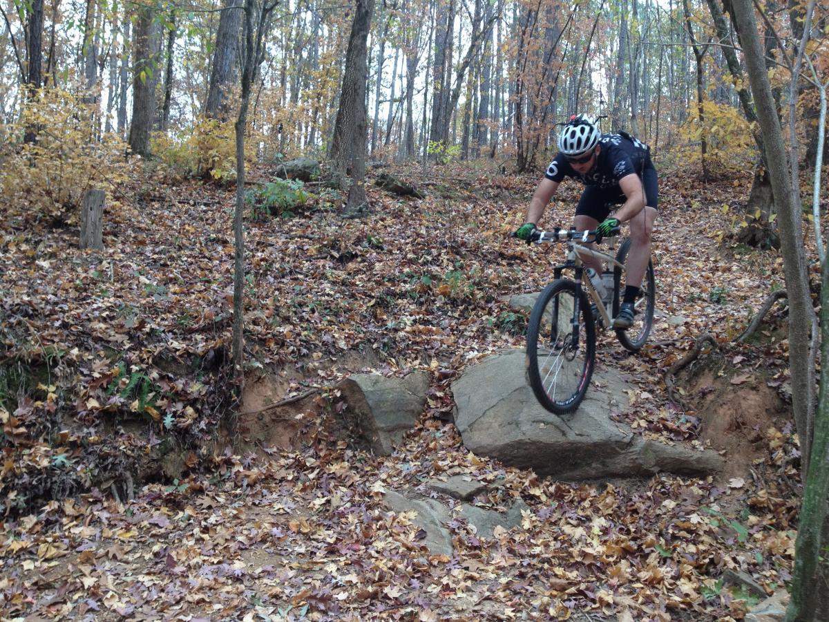 Vassago Jabberwocky: A mountain biker navigates over a rocky terrain in a wooded area during fall, surrounded by colorful autumn leaves and trees. The rider is airborne as they jump off a rock, demonstrating skill and control on the trail.