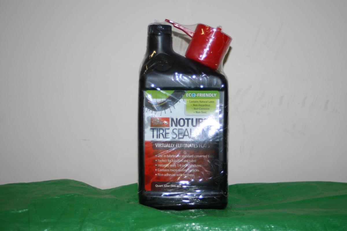 Stans Standard Kit: A bottle of eco-friendly tire sealant, labeled "Natural Tire Sealant," displayed against a light background. The bottle is black with a green and white label, indicating it contains natural latex and is non-toxic. It features a red cap, and the text highlights its ability to eliminate flats and its suitability for use in bicycles and tubes. The bottle is partially covered in plastic wrap. A green surface is visible at the bottom of the image.