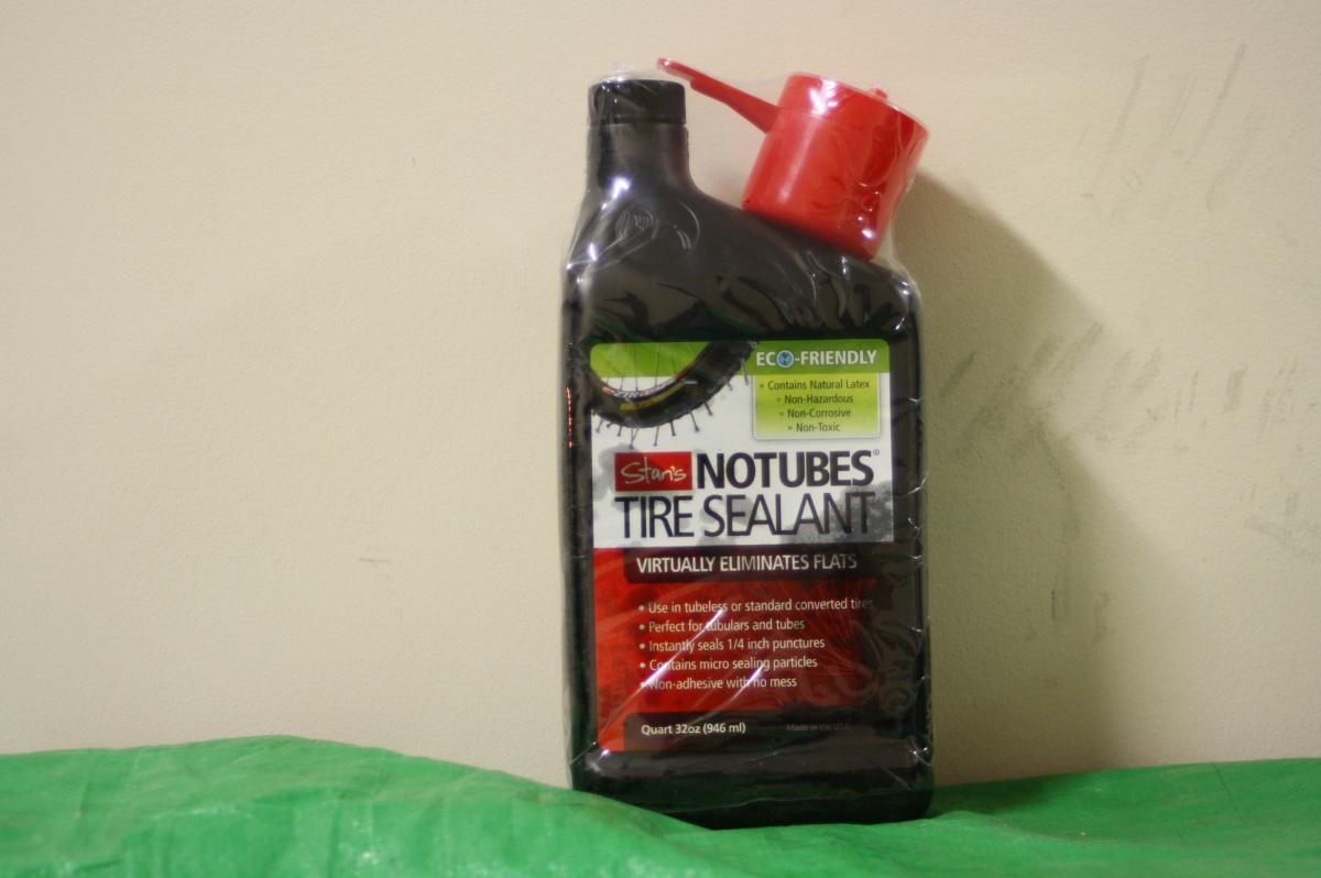 Stans Standard Kit: A 32-ounce bottle of Stan's NoTubes tire sealant, featuring a red dispensing nozzle and an eco-friendly label. The bottle is designed to help eliminate flat tires by instantly sealing punctures. It is placed on a green surface against a light-colored wall.