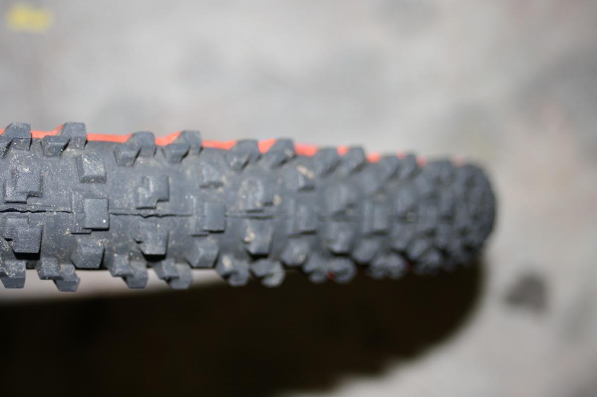 Panaracer Fire XC Pro: Close-up view of a bicycle tire showing a rugged tread pattern designed for off-road terrain, featuring a mix of square and rectangular lugs for enhanced traction. The tire is partially illuminated with a blurred background, highlighting its textures and details.