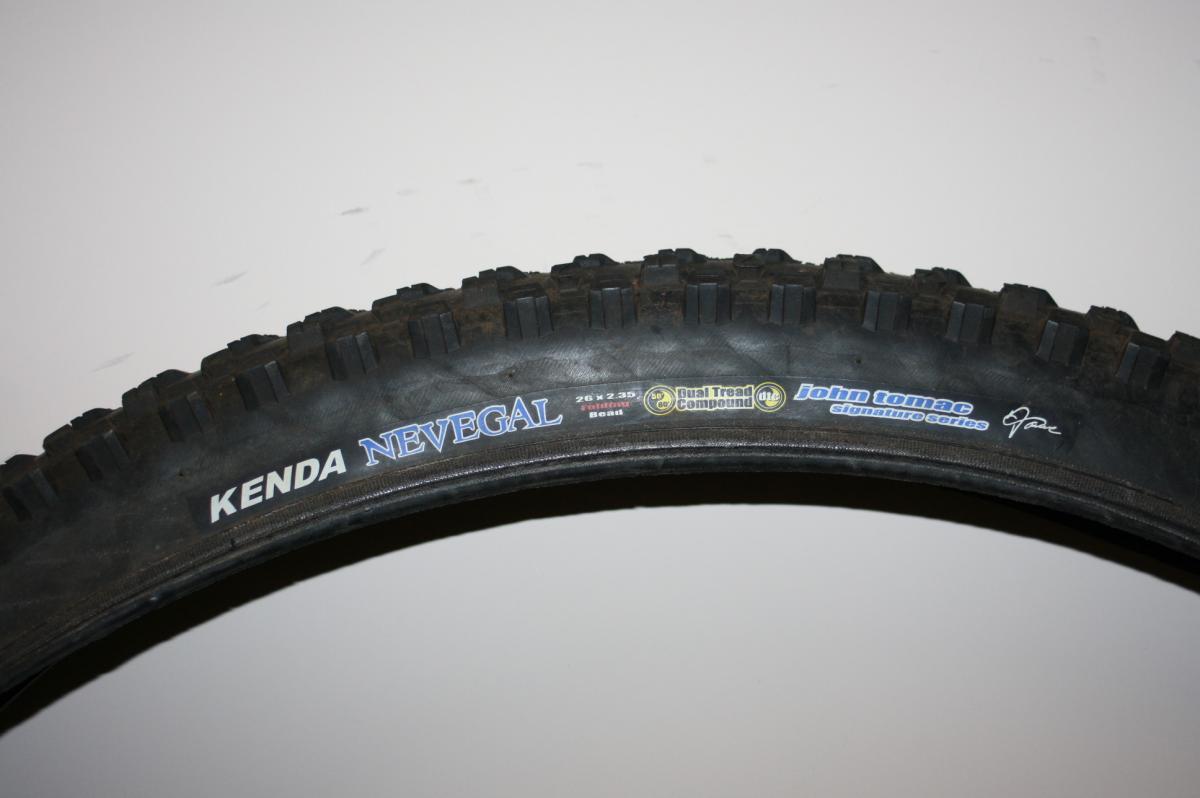 Kenda Nevegal: Alt text: Close-up view of a Kenda Nevegal mountain bike tire, displaying the brand name, model, and features such as 'Dual Tread Compound' and 'John Tomac Signature Series'. The tire size of 26 x 2.35 is also visible.